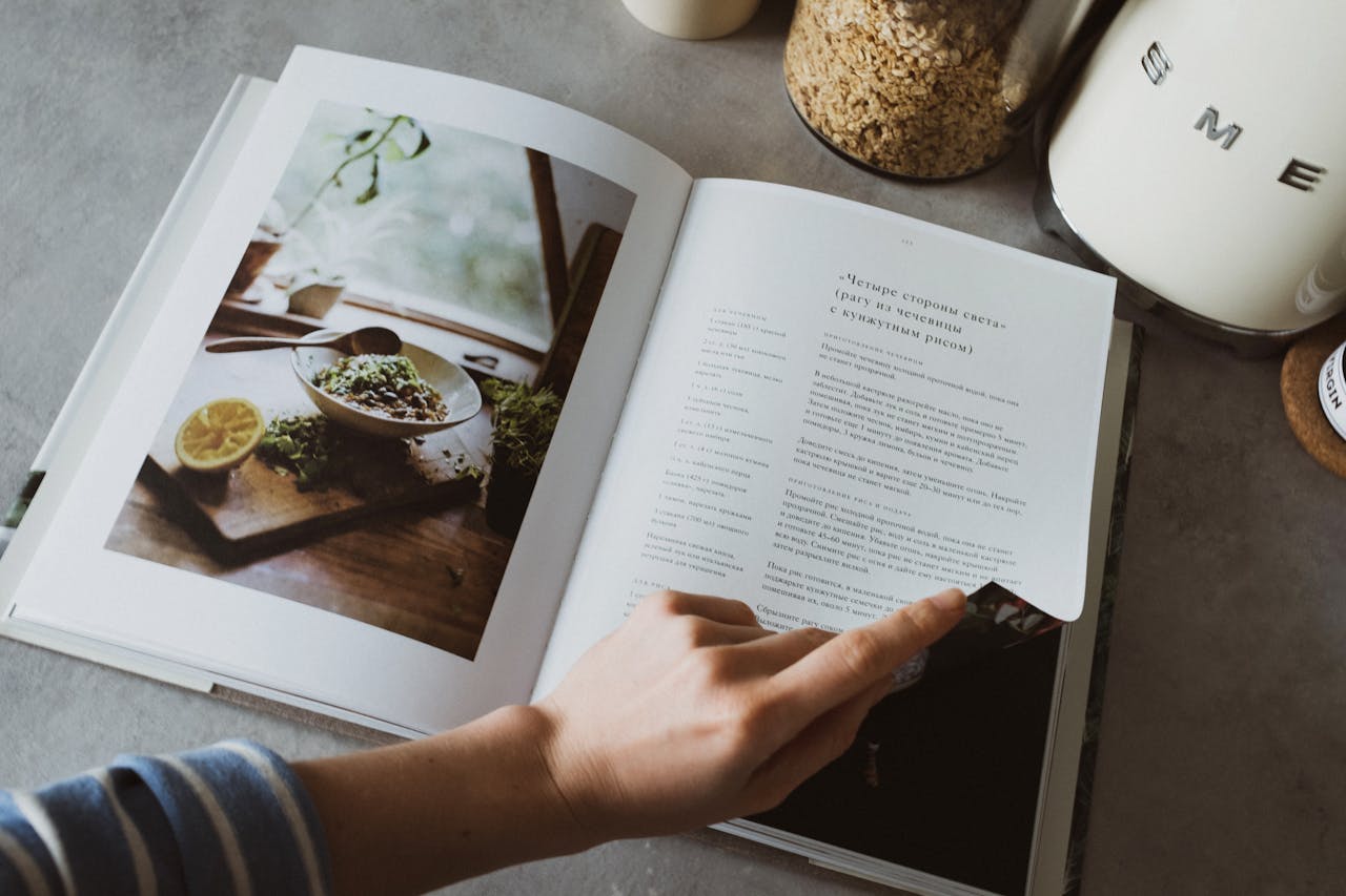 A person turns the page of a recipe book on a kitchen counter, surrounded by cooking ingredients.