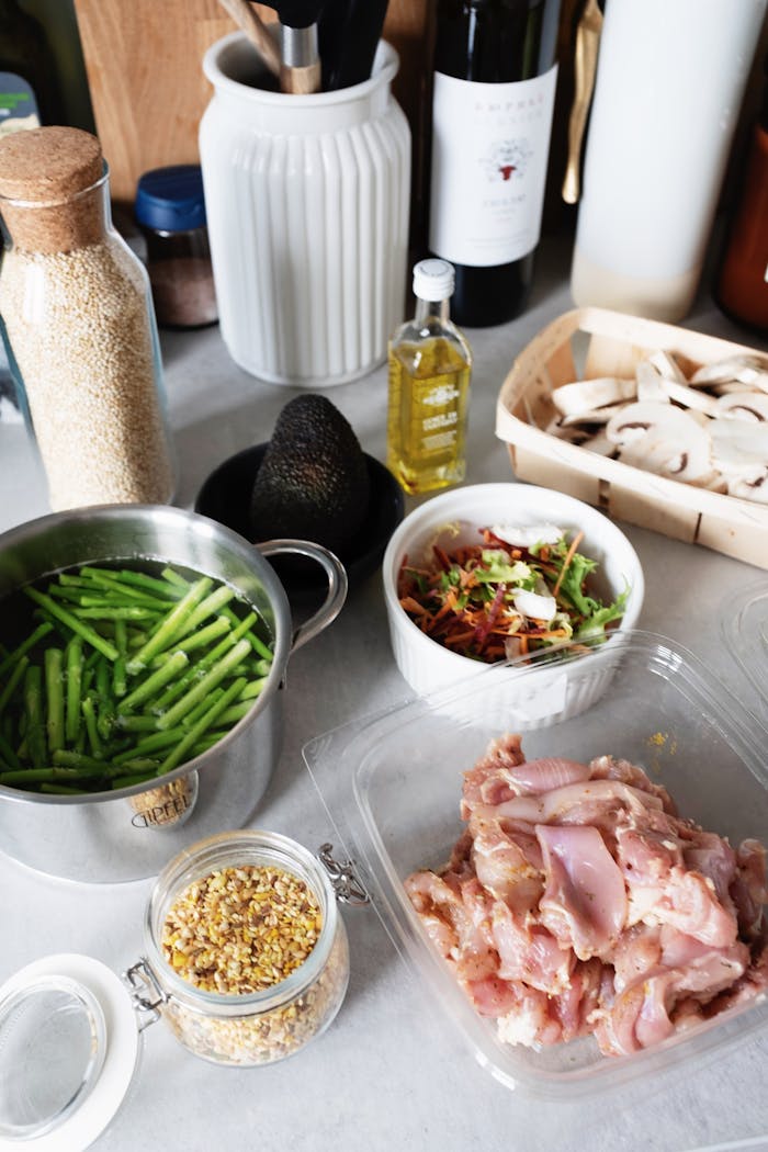 From above of raw chicken pieces in container green beans in pot of water and chopped champignons slices in wicker basket next to salad bowl avocado and spices in glass jar on countertop with various kitchen utensils